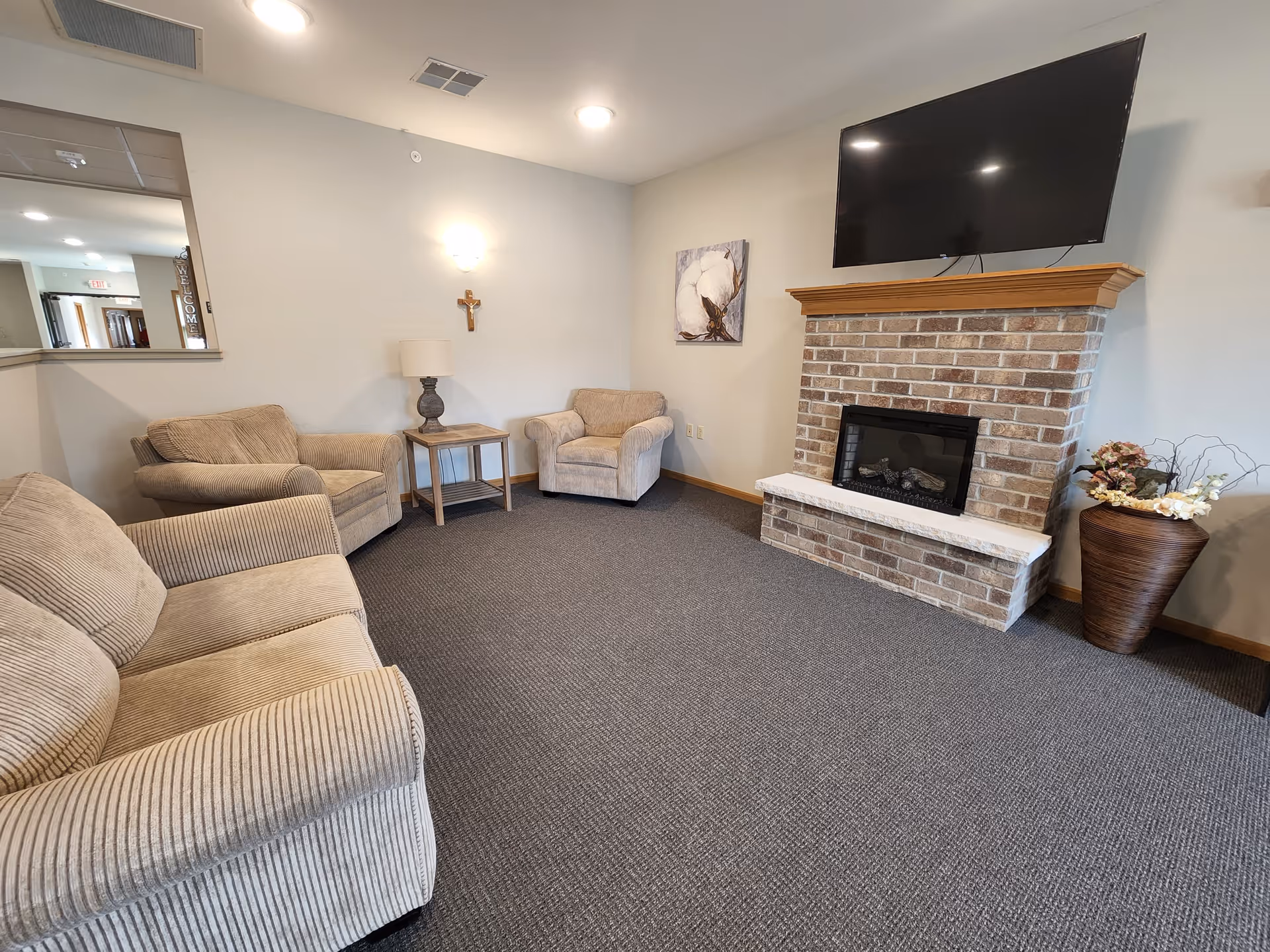 A cozy living room area with beige upholstered armchairs and a sofa arranged around a brick fireplace with a mounted flat-screen TV above it. There is a side table with a lamp between two chairs, a decorative cross on the wall, a floral painting, and a large vase with flowers on the floor next to the fireplace. The room has gray carpeting and light-colored walls.