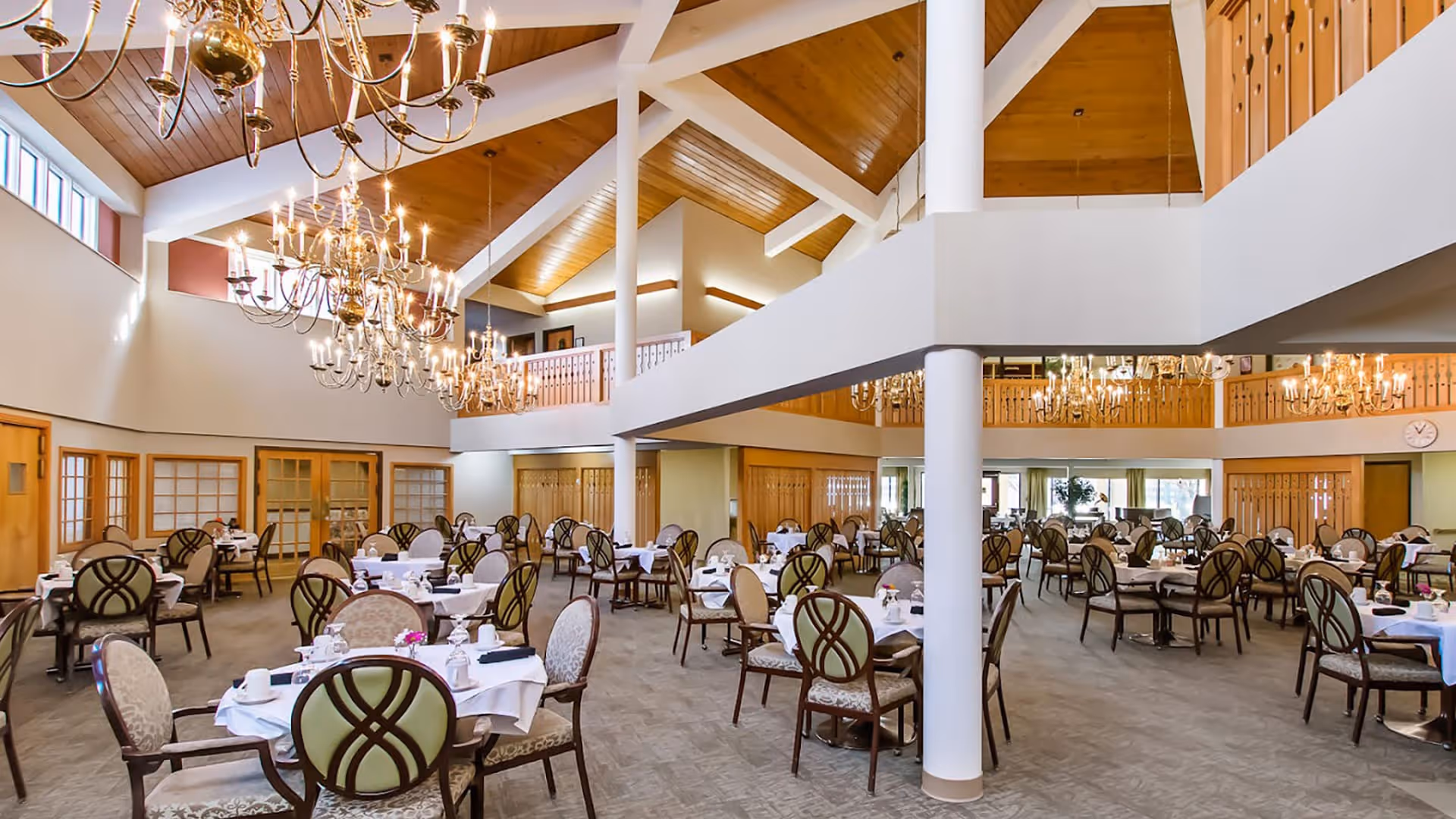 Spacious dining room with multiple round tables covered with white tablecloths, set with cups and plates. The room features high vaulted wooden ceilings with large chandeliers, beige walls, and a second-floor balcony overlooking the area. Chairs with patterned upholstery surround the tables.