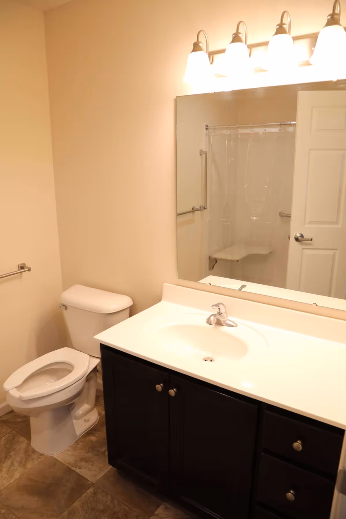 A bathroom with a white toilet, a dark brown vanity with a white sink, a large mirror above the sink, and a shower area with a clear plastic curtain and a built-in seat visible in the reflection.