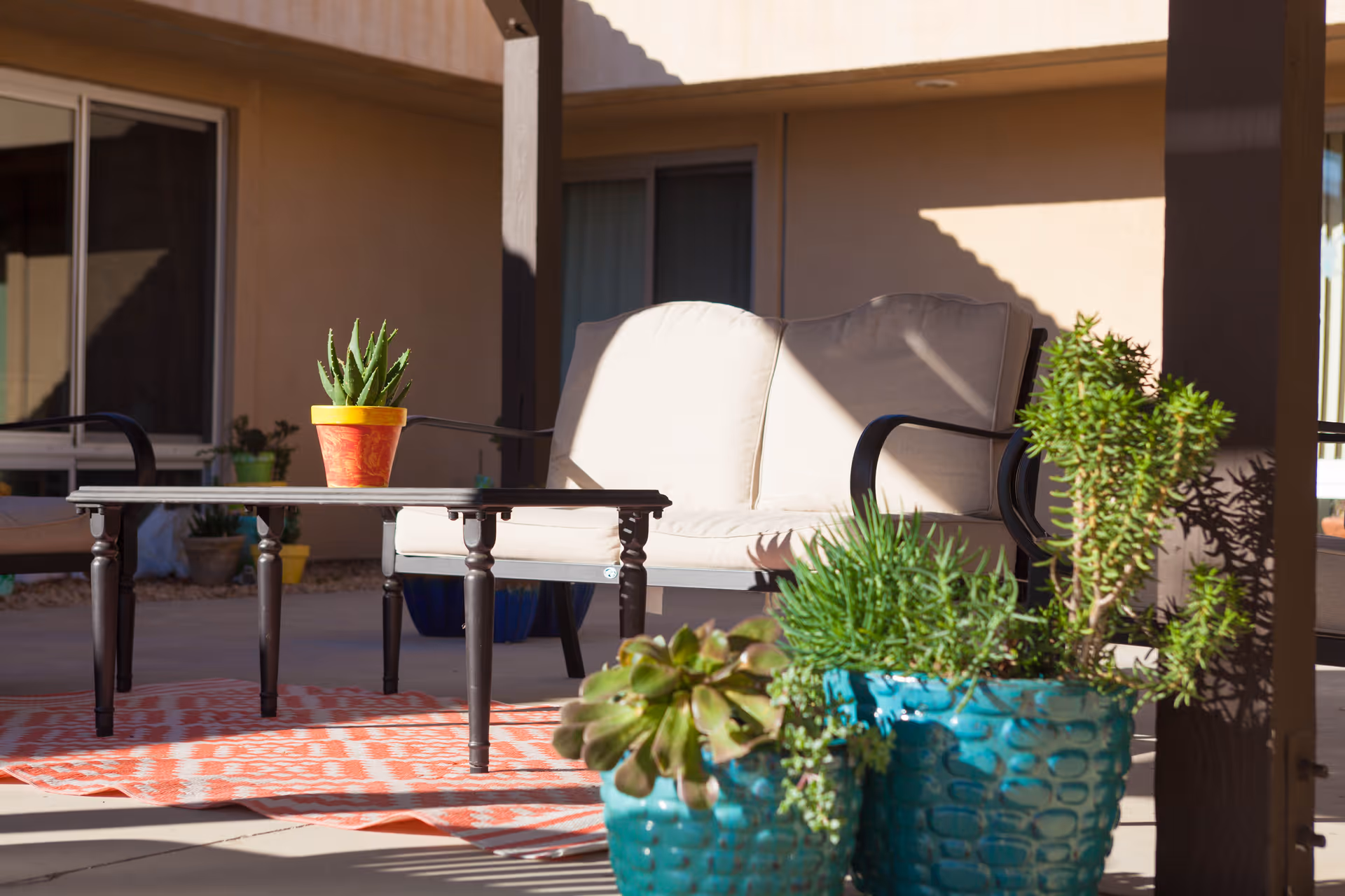 Sunlit outdoor patio with a cushioned loveseat, coffee table and several potted plants.