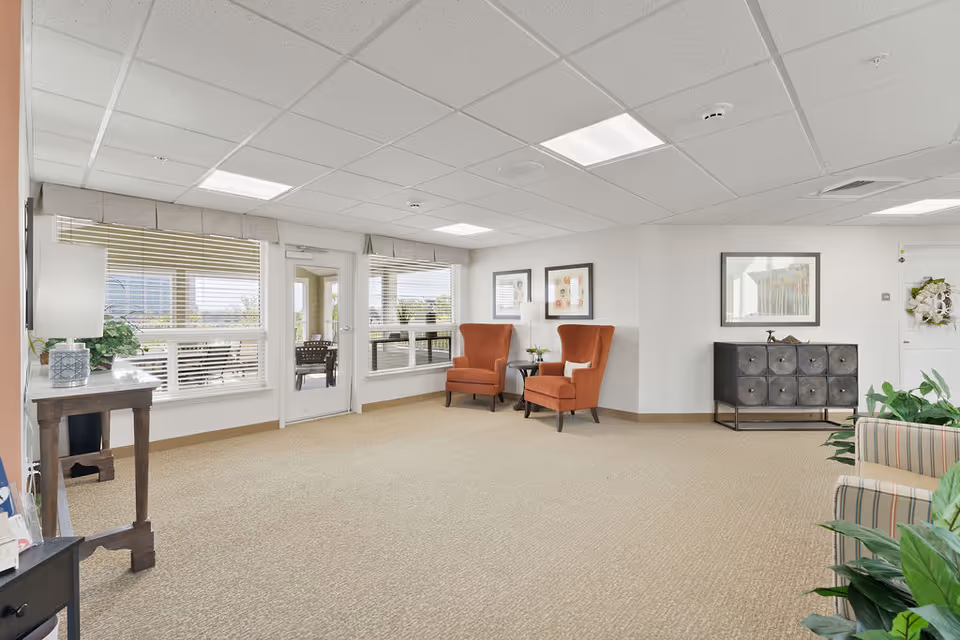 A bright and spacious common area with beige carpet, white walls, and a drop ceiling with fluorescent lighting. The room features two orange upholstered armchairs with a small round table between them, a black cabinet with decorative items on top, framed artwork on the walls, and several green plants. Large windows with blinds and a glass door provide natural light and a view of an outdoor patio area with seating.