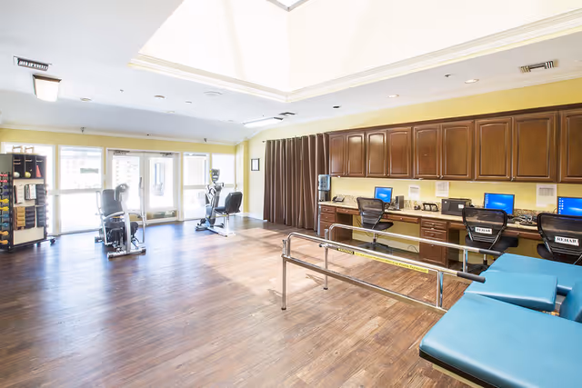 A spacious rehabilitation room with wooden flooring, exercise equipment including stationary bikes, a row of desks with computers and office chairs labeled 'REHAB', and padded treatment tables. The room has large windows allowing natural light and a high ceiling with a skylight.