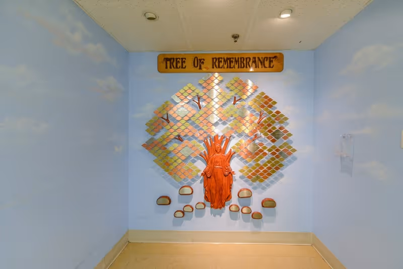 A wall display titled 'Tree of Remembrance' featuring a wooden sculpture of a figure with outstretched arms in front of a stylized tree made of small, colorful plaques on a light blue wall with painted clouds.