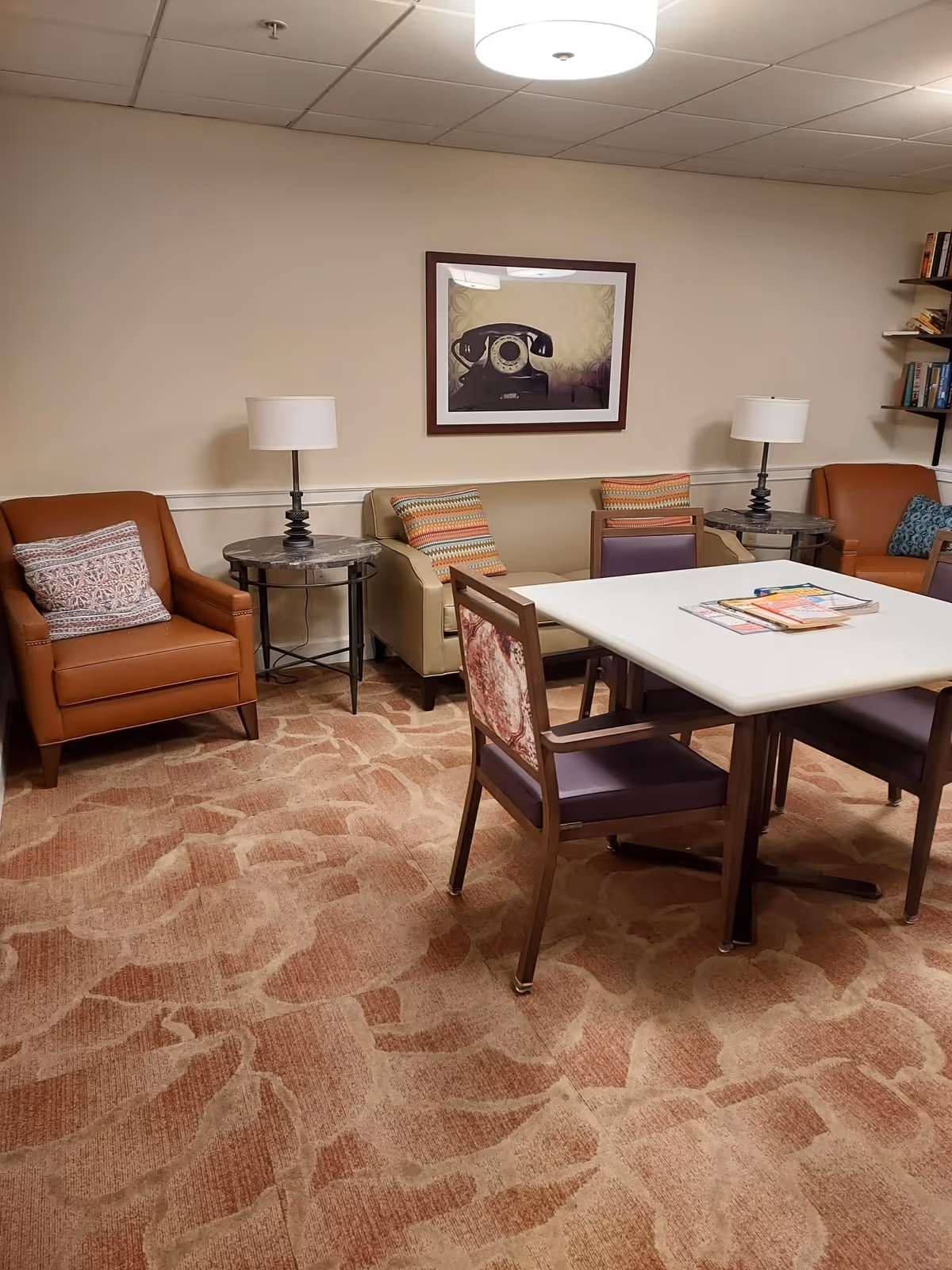 A cozy senior living common area with a beige sofa, two brown armchairs, and a white square table surrounded by four chairs. The room has a patterned carpet, two side tables with lamps, a framed picture of a vintage telephone on the wall, and bookshelves with books on the right side.