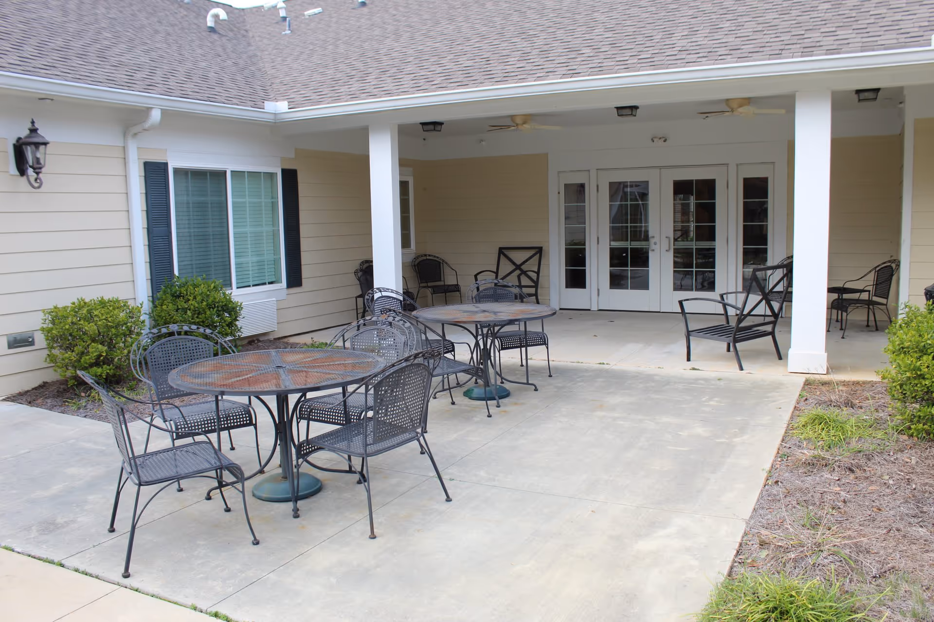Outdoor patio area at The Summit of Atmore Assisted Living Facility with metal tables and chairs on a concrete surface, beige siding walls, white pillars, and glass doors leading inside.
