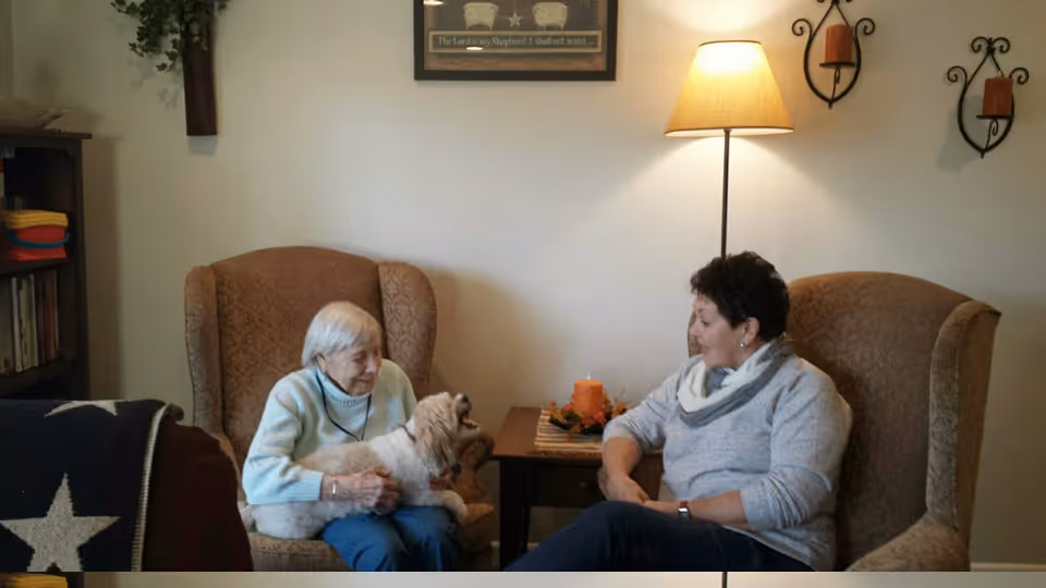 Two women sit in armchairs in a cozy living room, one holding a small dog while they converse.