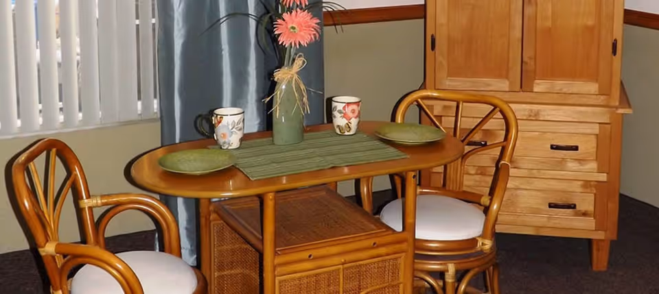 Small wooden dining table with two chairs, place settings, a vase with a flower, and a wooden cabinet in a room.