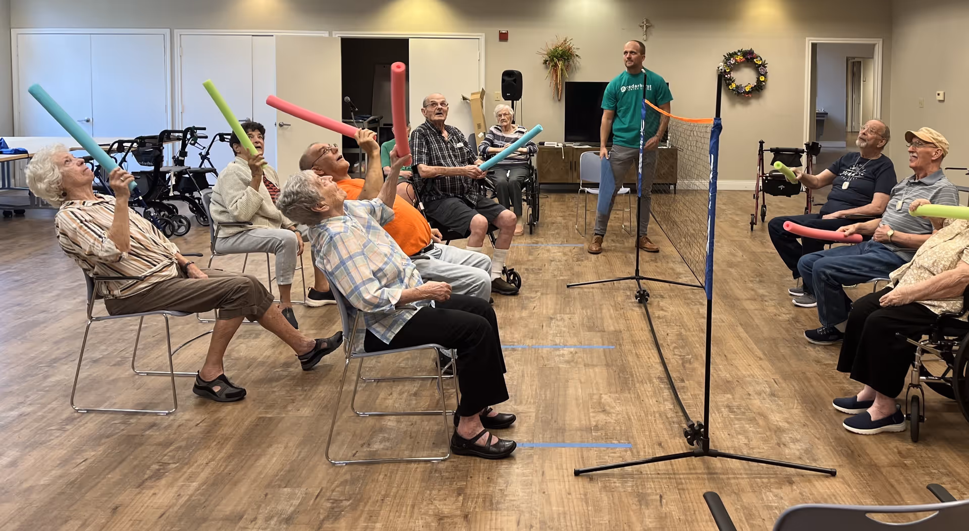 A group of elderly people seated in chairs in a spacious room, playing a seated game with colorful foam noodles and a small net in the center. A staff member stands near the net, overseeing the activity. The room has wooden flooring and light-colored walls with minimal decorations.