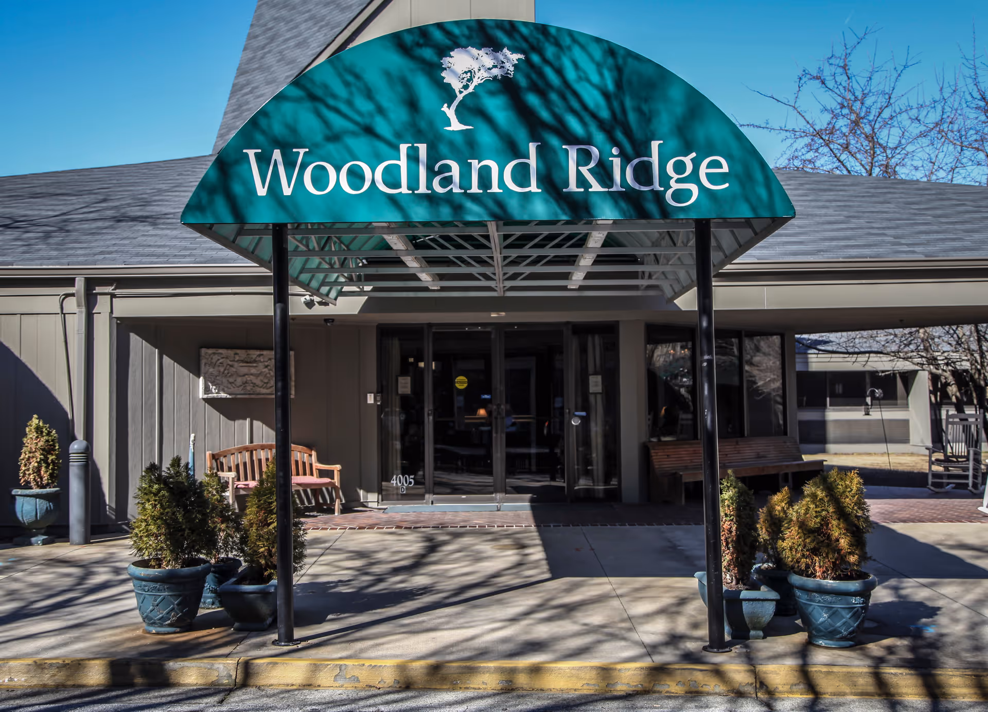Entrance to Woodland Ridge Assisted Living facility with a green awning displaying the facility name and a tree logo. There are potted plants and benches near the entrance under a clear blue sky.