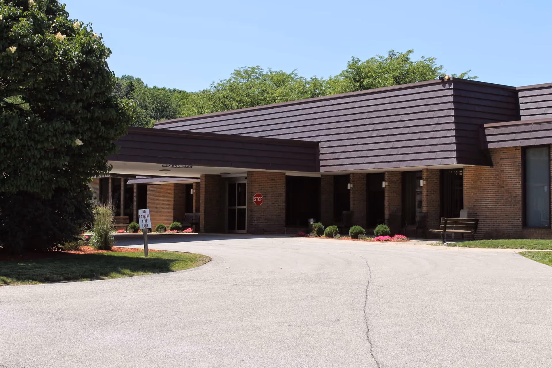 Exterior view of a single-story brick building with a dark brown roof, a covered entrance, a stop sign, and a 'No Parking Fire Lane' sign. There are bushes, flowers, and a bench near the entrance, with trees and a clear blue sky in the background.