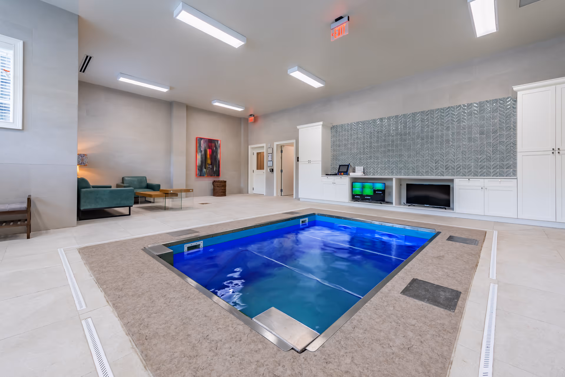 Indoor therapy pool with blue water in a modern common room featuring seating, cabinets, and artwork.