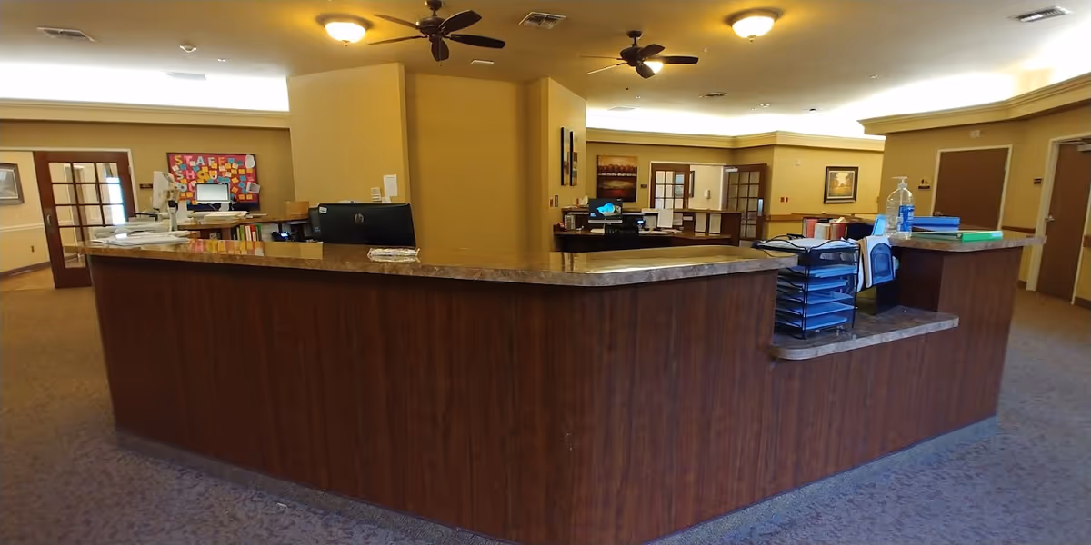Reception area inside Onion Creek Nursing and Rehabilitation Center with a large wooden front desk, computer monitors, office supplies, ceiling fans, and beige walls with framed artwork.