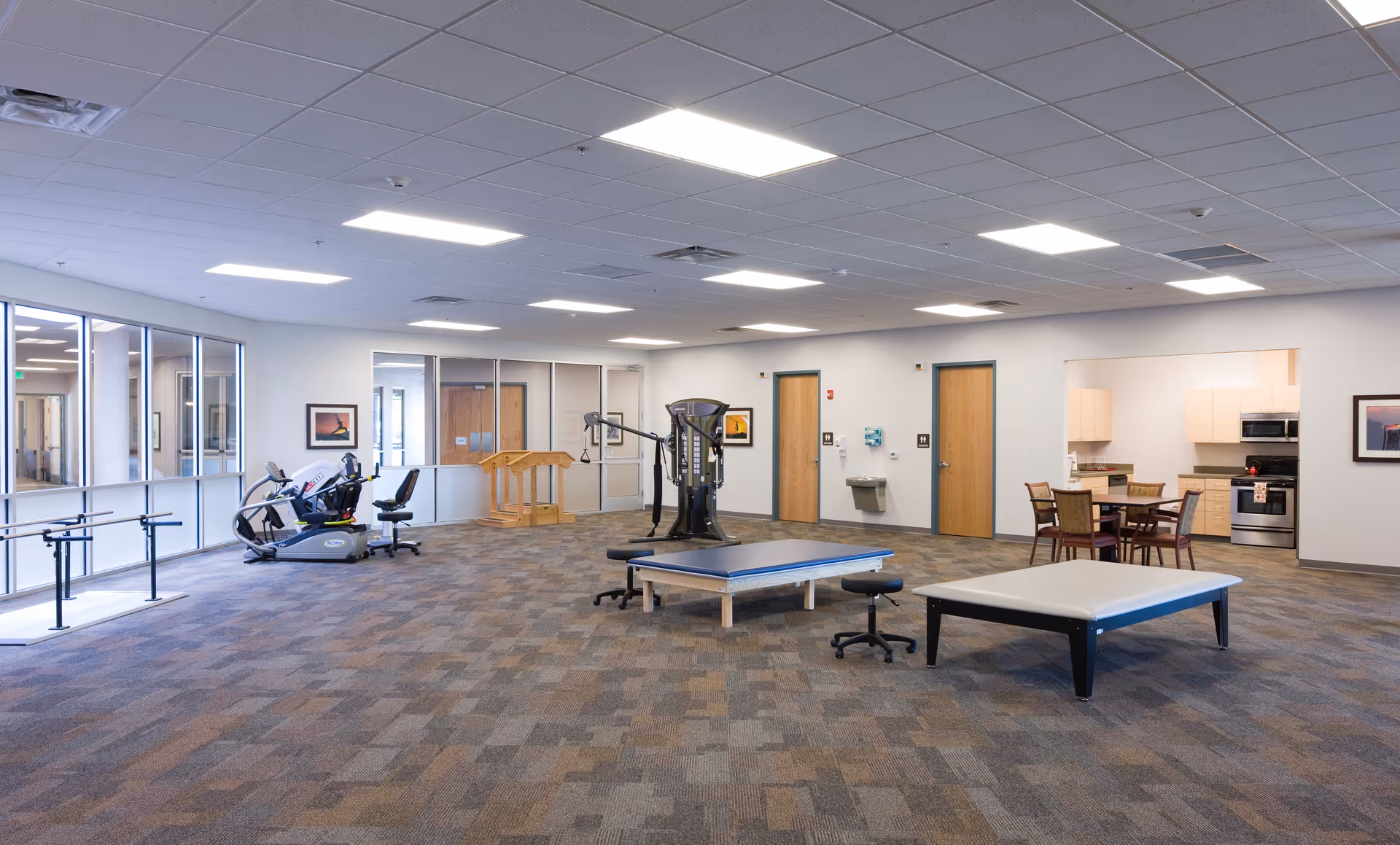A spacious rehabilitation or therapy room with exercise equipment including a recumbent bike, a multi-function exercise machine, two therapy tables, and a small dining area with a table and chairs near a kitchenette. The room has carpeted floors, white walls, and a drop ceiling with fluorescent lighting. There are several doors and windows along the walls.