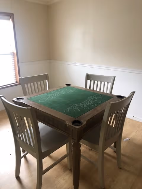 A small square wooden table with a green felt top designed for card games, surrounded by four white wooden chairs, placed in a room with light-colored wooden flooring and a window with blinds on the left side.