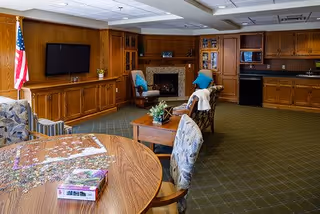 A cozy common area in a senior living facility featuring a round wooden table with a partially completed puzzle and puzzle box on it, upholstered chairs, a fireplace with a mantle, a flat-screen TV mounted on a wooden paneled wall, and a kitchenette with wooden cabinets and appliances. An American flag is displayed near a window with curtains.