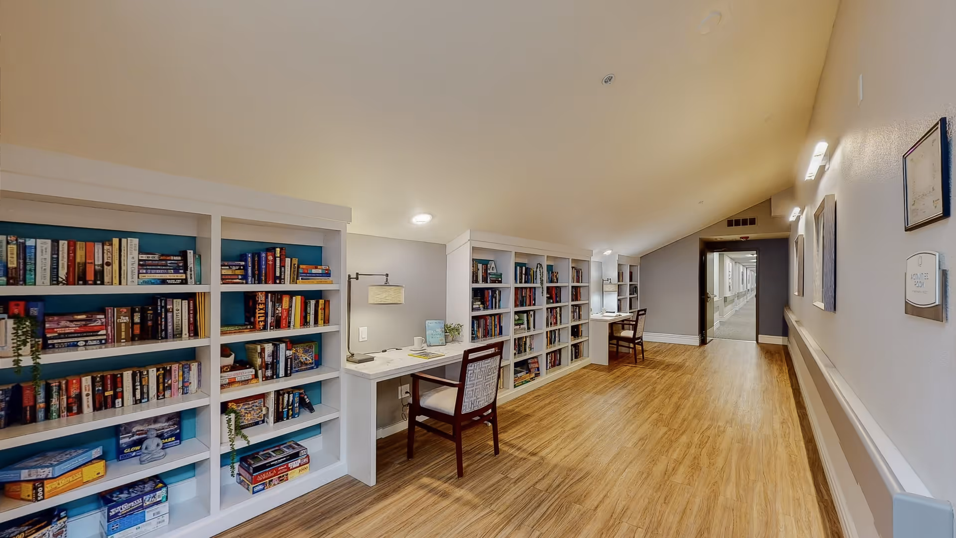 A well-lit hallway in a senior living facility featuring built-in white bookshelves filled with books and board games. There are two desks with chairs and lamps integrated into the bookshelves along the left side. The floor is wood, and the walls are painted light gray with some framed artwork and a thermostat on the right wall. The hallway extends into the distance with additional lighting and a doorway at the end.