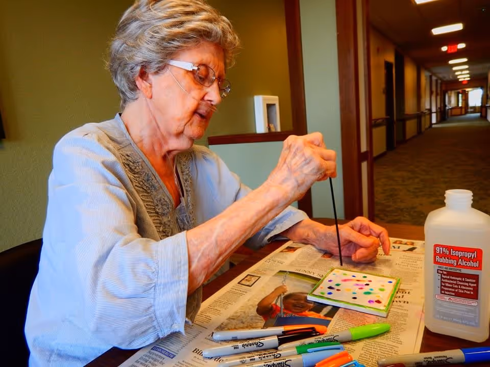 An elderly woman wearing glasses and a light blue blouse is sitting at a table in a hallway, painting on a small square canvas with a thin brush. The table is covered with newspaper and various colored markers are scattered around. A bottle of 91% isopropyl rubbing alcohol is also on the table.