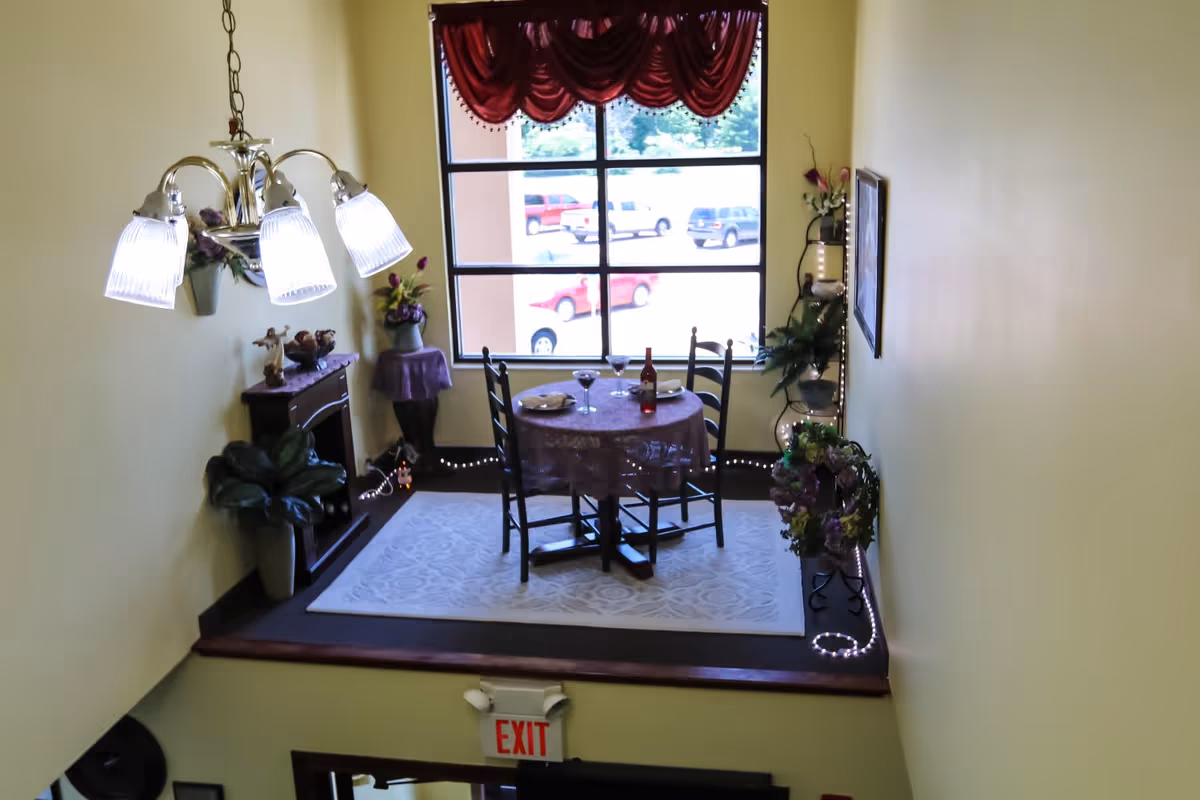 Small staged dining nook with a round table set for two, a chandelier, potted plants, decorative accents and a large window above an EXIT sign.
