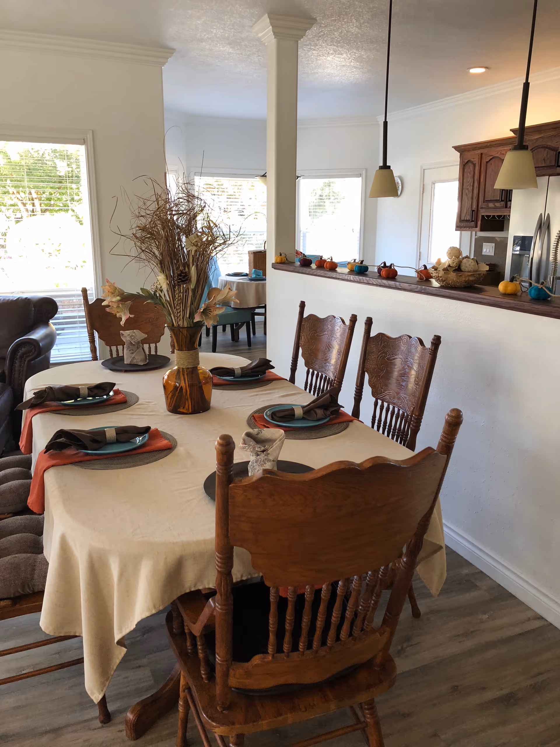 A dining room with a wooden table covered by a beige tablecloth set for six people with plates, napkins, and a centerpiece vase with dried flowers. The room has wooden chairs around the table, a partial view of a living area with a leather couch, and a kitchen with wooden cabinets and pendant lights in the background. Small decorative pumpkins are placed on the kitchen counter.
