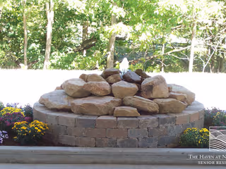 A circular stone water fountain with a pile of large rocks in the center, surrounded by colorful flowers and greenery in the background.