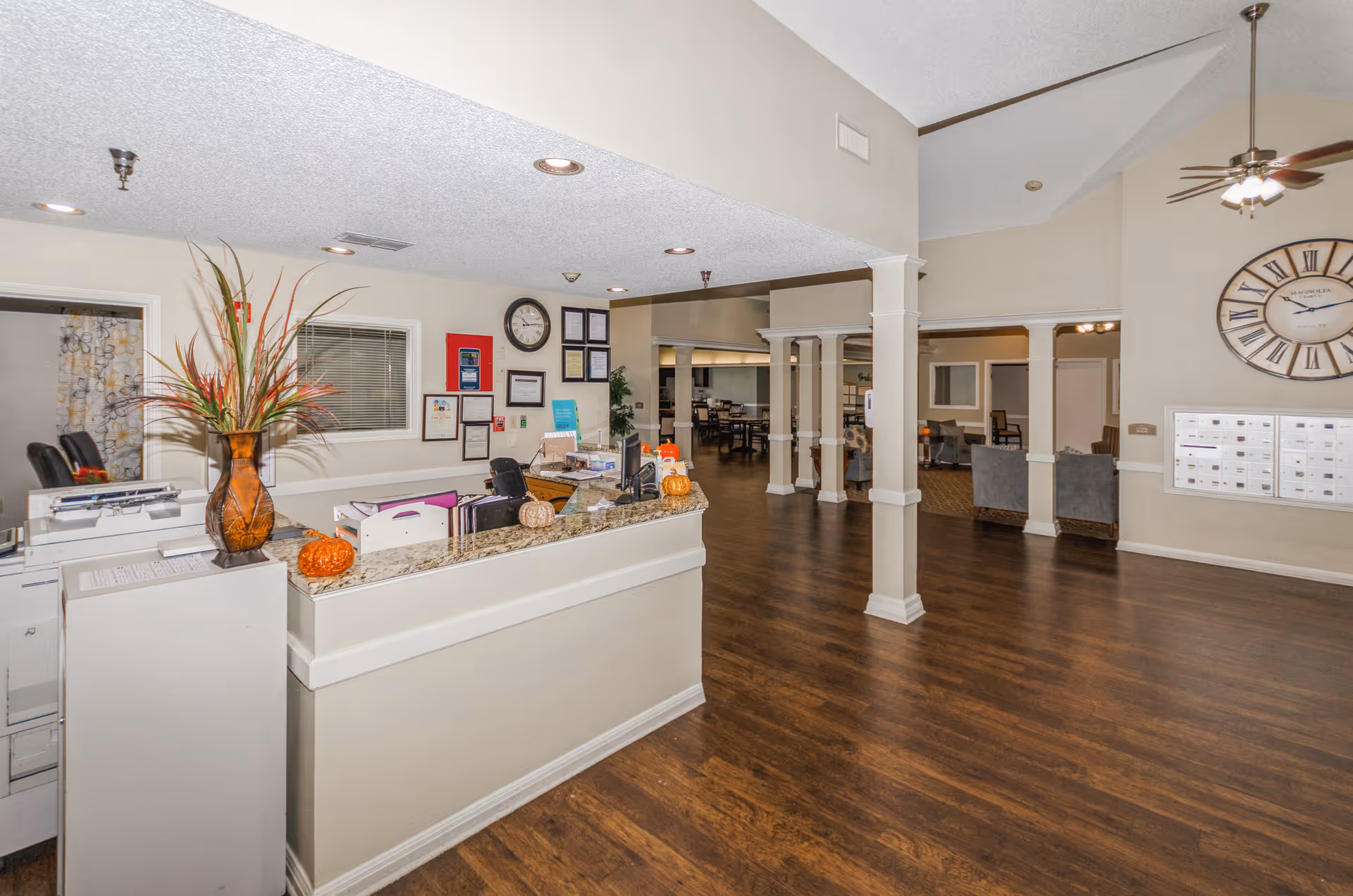 Interior view of a senior living facility reception area with a front desk decorated with a vase of tall plants and small pumpkins. Behind the desk are framed certificates and a clock on the wall. The space has wooden flooring, beige walls, and an open layout leading to a dining area and a lounge with chairs. A large wall clock and mailboxes are visible on the right side.
