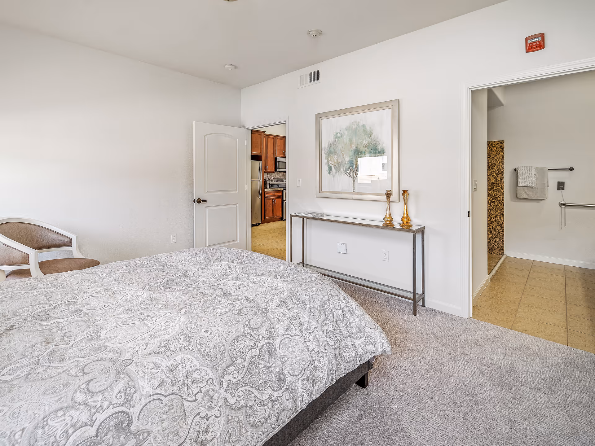 A bedroom with a bed covered in a patterned gray and white comforter, a beige and white chair in the corner, and a narrow console table against the wall with two decorative vases and a framed painting of a tree above it. The room has light-colored walls and carpeted floor. Two doorways are visible: one leading to a kitchen with wooden cabinets and stainless steel appliances, and the other leading to a bathroom with tiled floor and a towel rack.