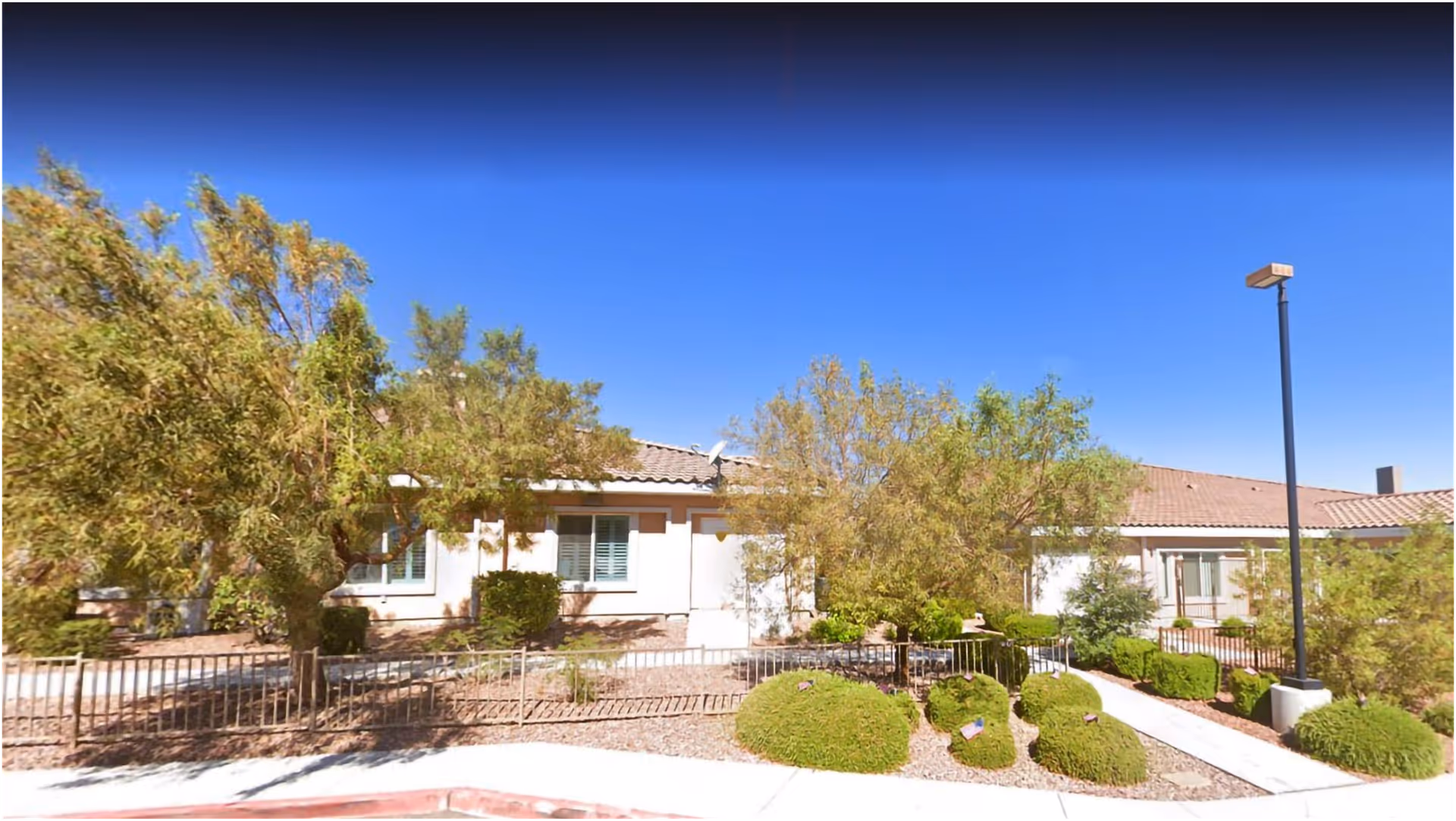 Exterior view of a single-story building with a tiled roof, surrounded by trees, bushes, and a metal fence under a clear blue sky.
