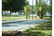 Shuffleboard courts with benches on a grassy, tree-lined outdoor area near residential buildings.
