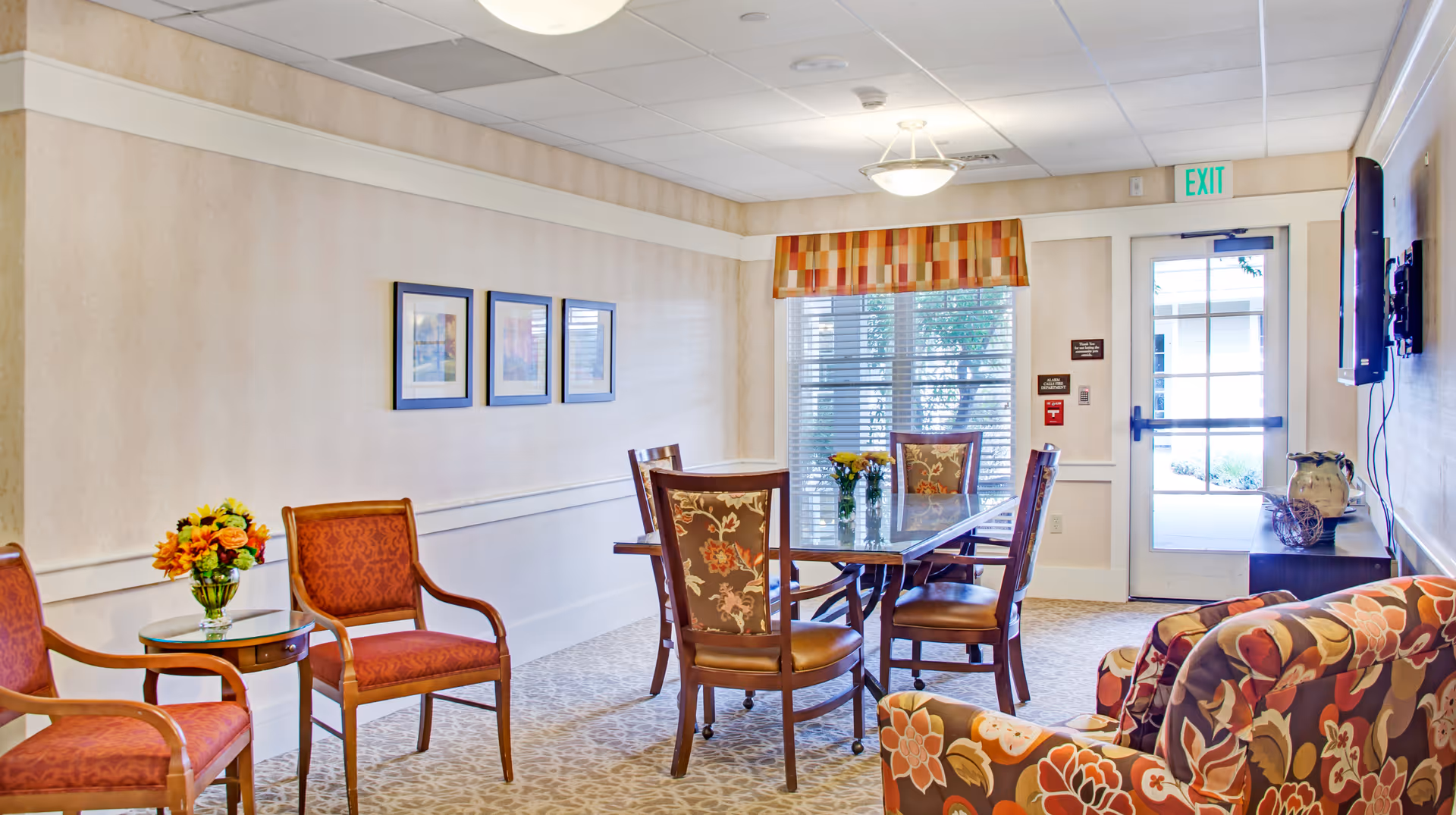 A bright and cozy common area in a senior living facility featuring a glass-top dining table with six chairs, two upholstered armchairs with floral patterns, two red cushioned chairs, a small round side table with a flower vase, framed artwork on the wall, and a door with an exit sign leading outside.