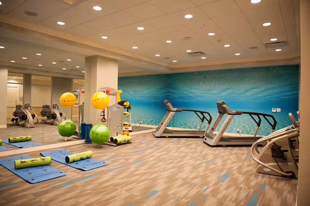 Bright indoor fitness room with treadmills, exercise mats, stability balls, and a mirrored wall featuring an ocean mural.