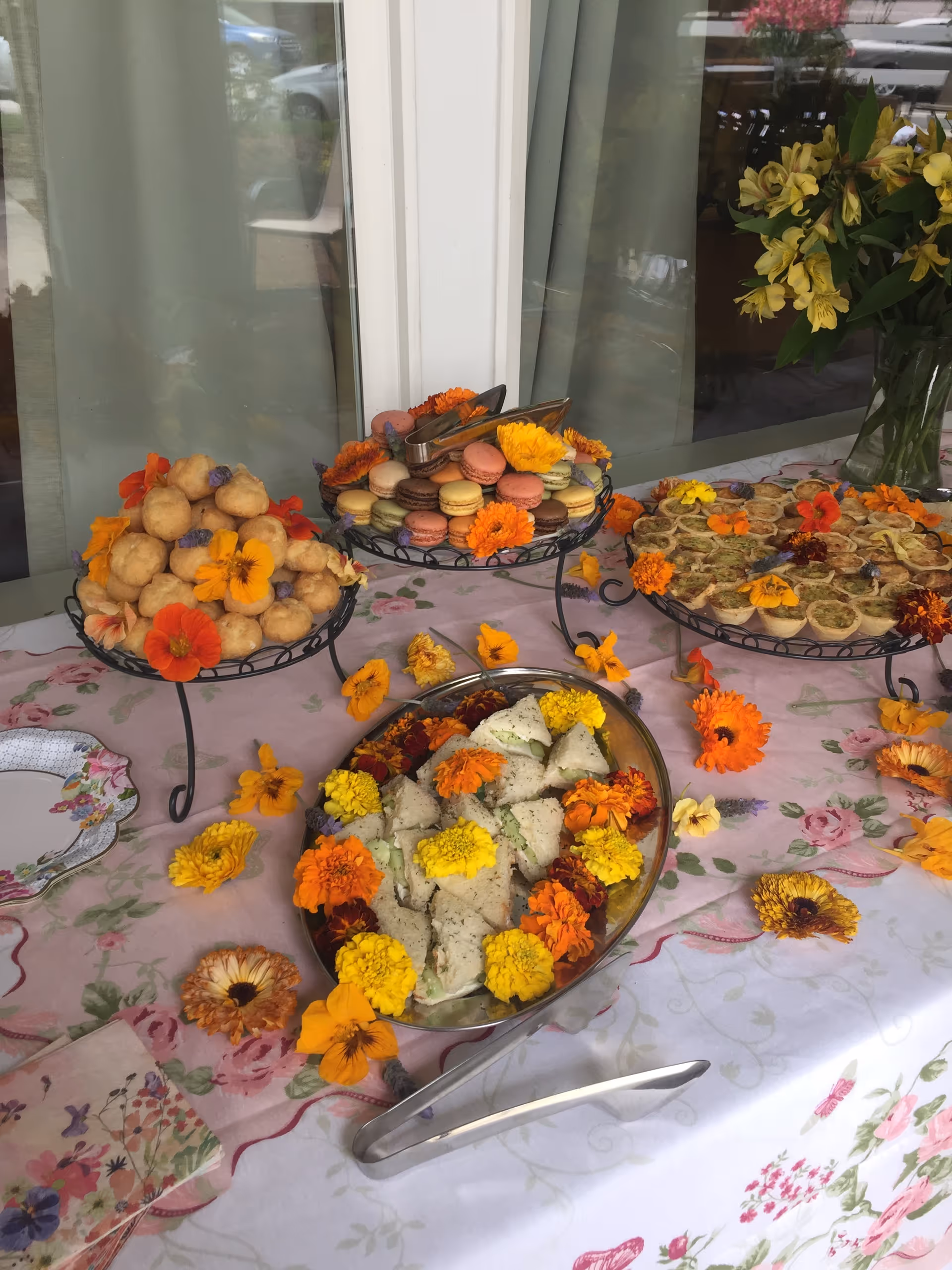 Table arranged with assorted pastries, macarons and finger sandwiches decorated with colorful edible flowers on tiered serving stands.