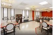 A bright dining room with multiple tables and chairs arranged neatly. The room has large windows with blinds, a bookshelf, and red accent walls. The ceiling has recessed lighting and decorative chandeliers.