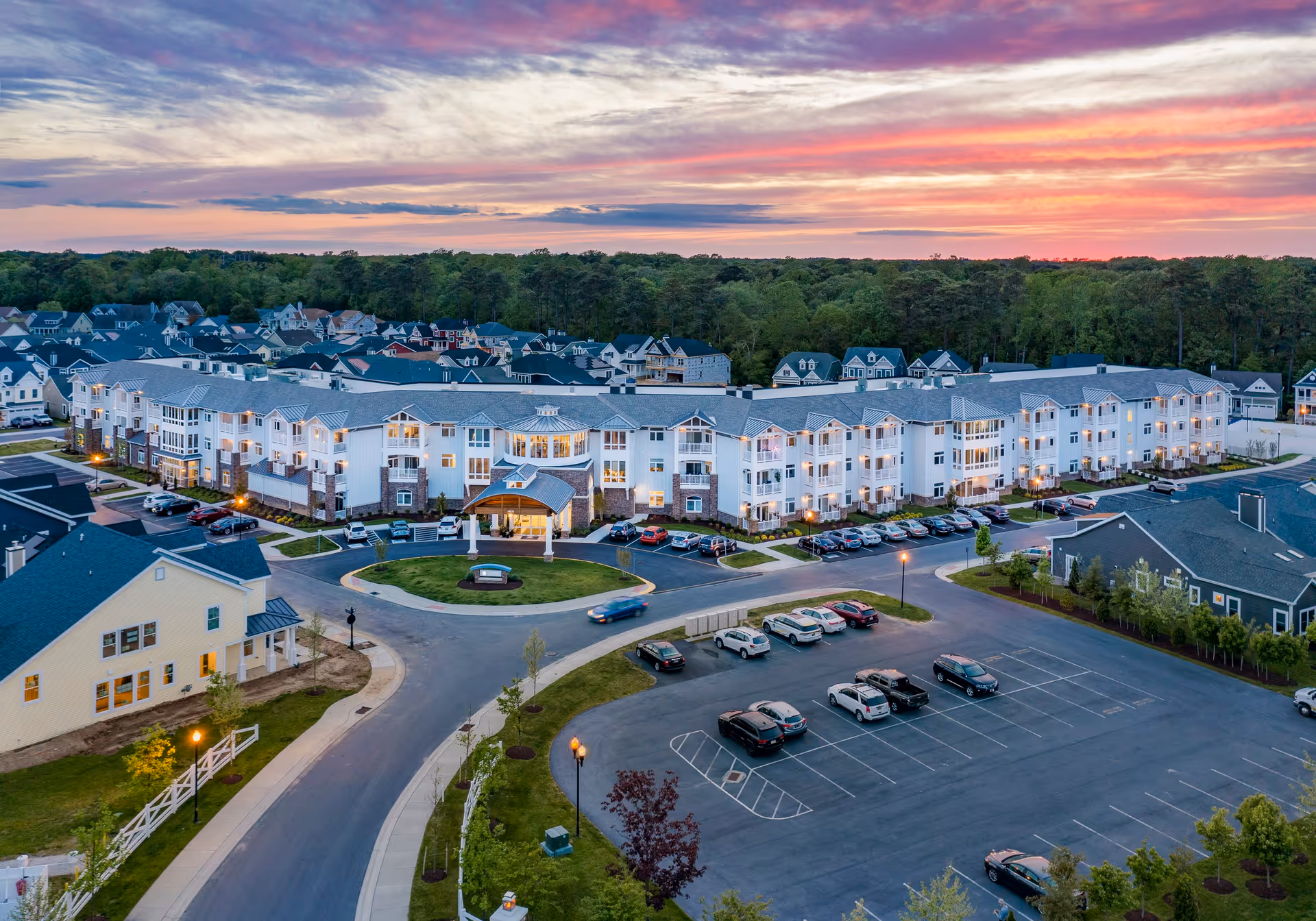 Aerial view of The Lodge at Truitt Homestead senior living facility at sunset, showing a large multi-story building with many windows and balconies, surrounded by parking lots with cars, landscaped greenery, and a neighborhood of houses in the background under a colorful sky.