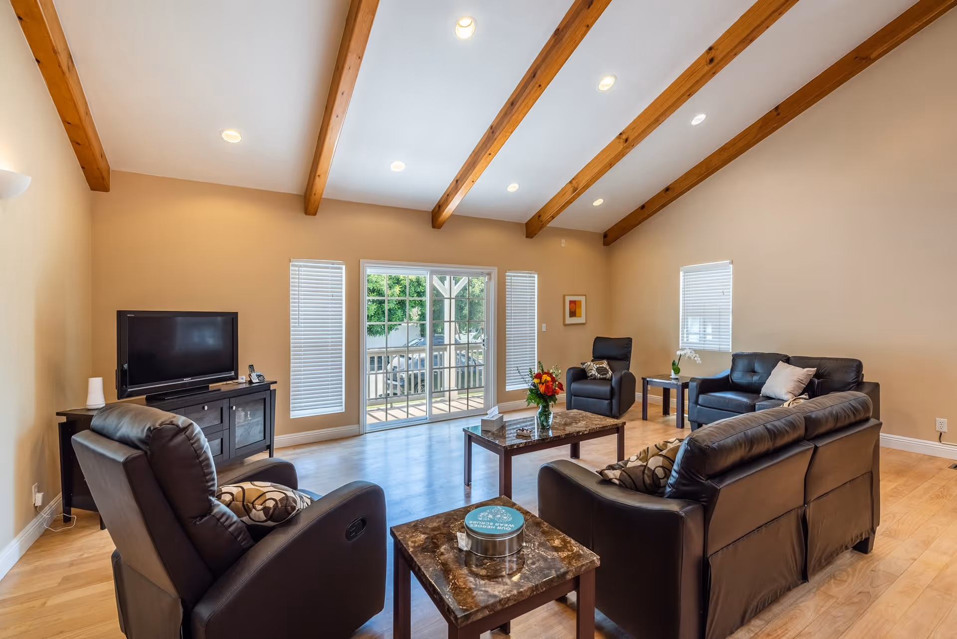 A spacious living room with wooden ceiling beams, recessed lighting, and light-colored walls. The room features a black leather sofa, two black leather armchairs, a TV on a black stand, and two marble-top coffee tables. Large sliding glass doors and windows with blinds allow natural light to enter the room.
