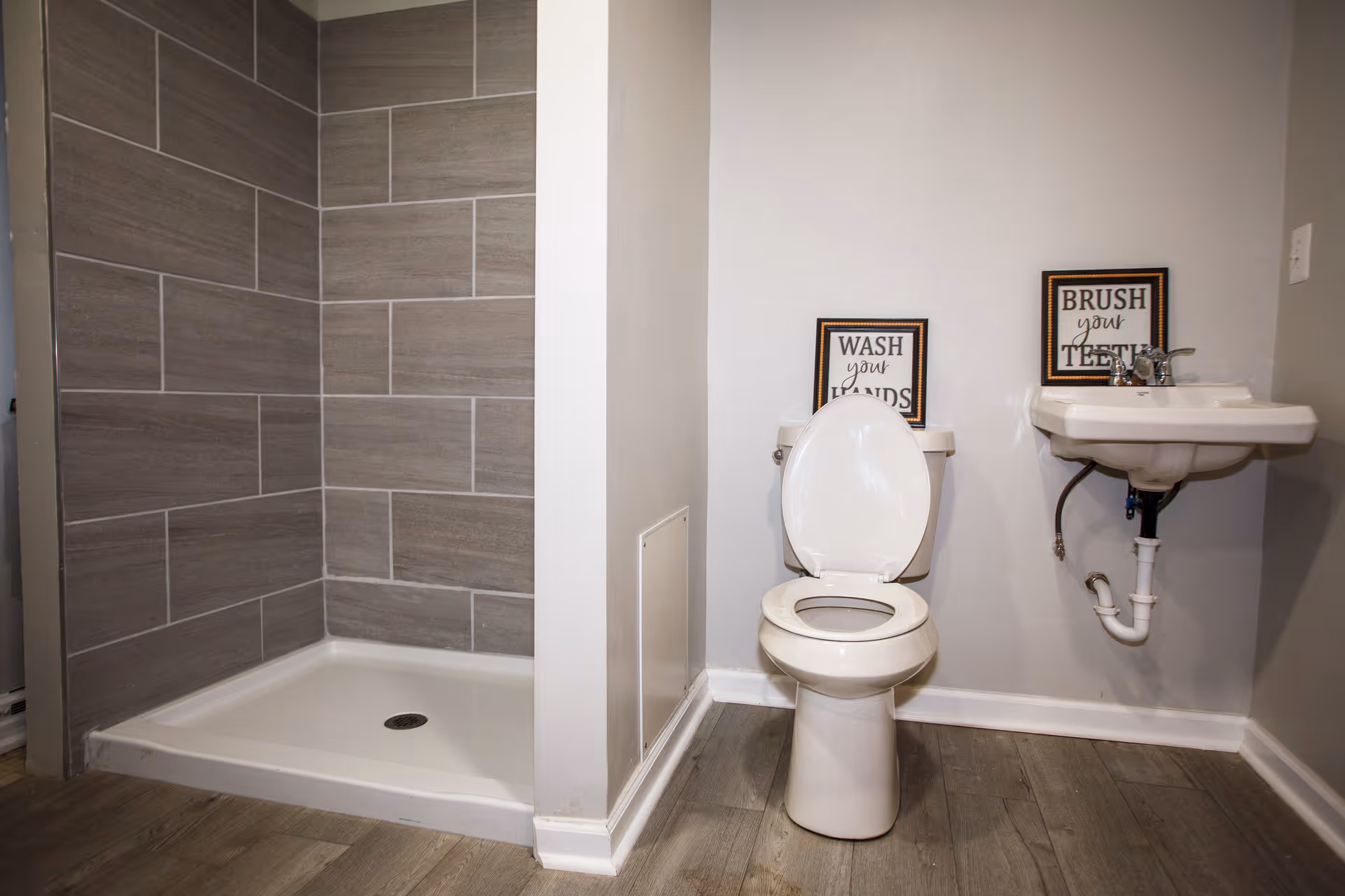 Bathroom with a gray-tiled walk-in shower on the left, a toilet in the center, and a wall-mounted sink with framed signs on the right.