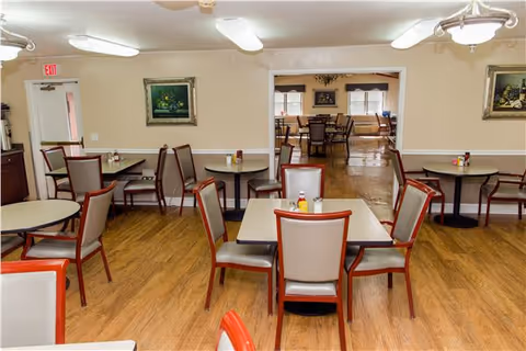 Interior view of a dining room in a senior living facility with several tables and chairs arranged on a wooden floor. The walls are beige with white trim, decorated with framed paintings. There is an open doorway leading to another room with more tables and chairs, and windows letting in natural light.