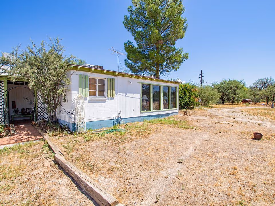 Exterior view of a single-story white building with green shutters and large windows, surrounded by dry grass and sparse vegetation under a clear blue sky.