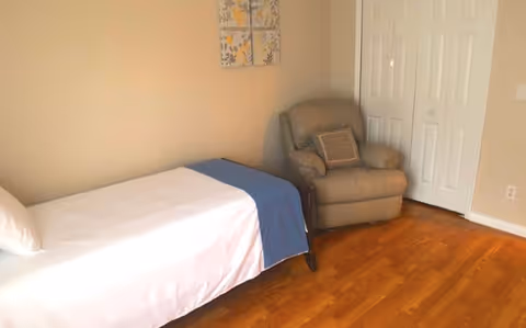 A simple bedroom with a single bed covered in white sheets and a blue blanket at the foot. Next to the bed is a beige recliner chair. The room has wooden flooring, beige walls, a white closet door, and a decorative wall hanging with floral patterns.