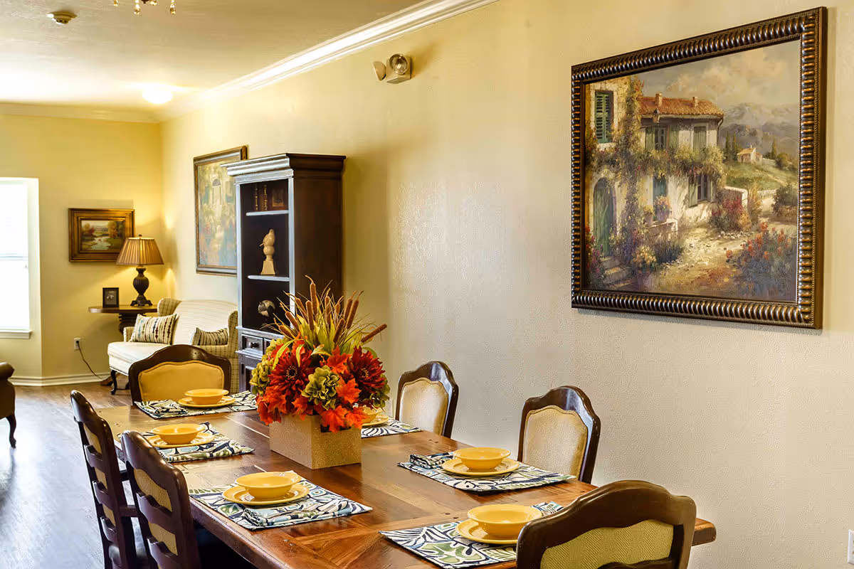 A warmly lit dining area with a wooden table set for six people, featuring yellow bowls and patterned placemats. A centerpiece with red and green flowers decorates the table. In the background, there is a beige sofa, a dark wooden cabinet with decorative items, a side table with a lamp, and framed paintings on the walls.