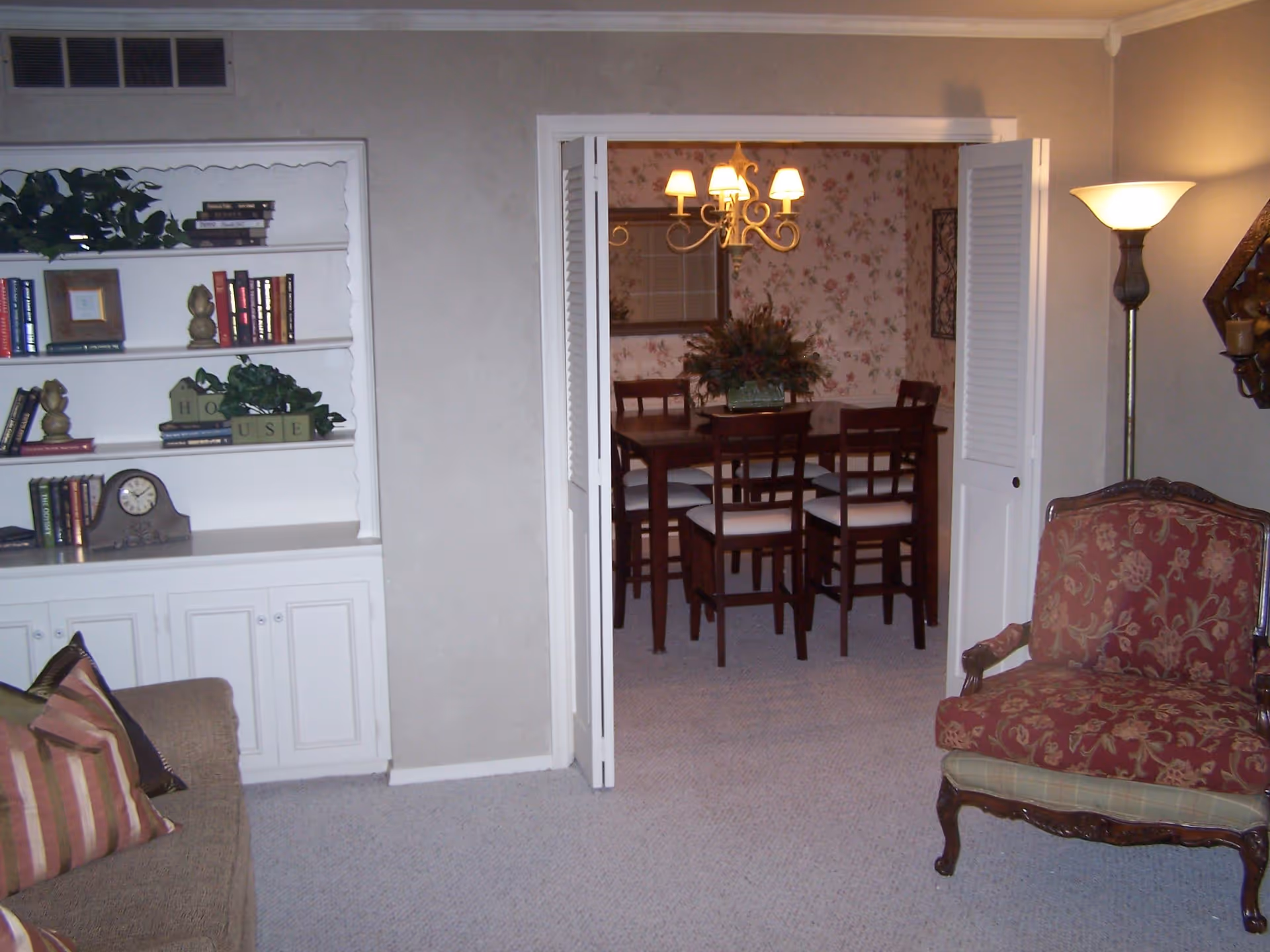 Interior view of a living room with a beige couch with striped pillows on the left, a white built-in bookshelf with books and decorative items, and a red patterned armchair on the right. In the background, through open white folding doors, there is a dining room with a wooden dining table, six chairs with white cushions, a floral wallpaper, and a chandelier with five lamps.