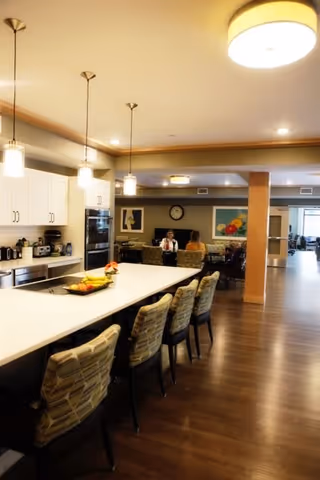 A spacious and well-lit kitchen and dining area in a senior living facility. The kitchen features a long white island with four cushioned chairs, pendant lights hanging above, and modern appliances along the back wall. In the background, two people are seated at a table in a lounge area with artwork and a clock on the wall. The floor is wooden, and the overall atmosphere is warm and inviting.