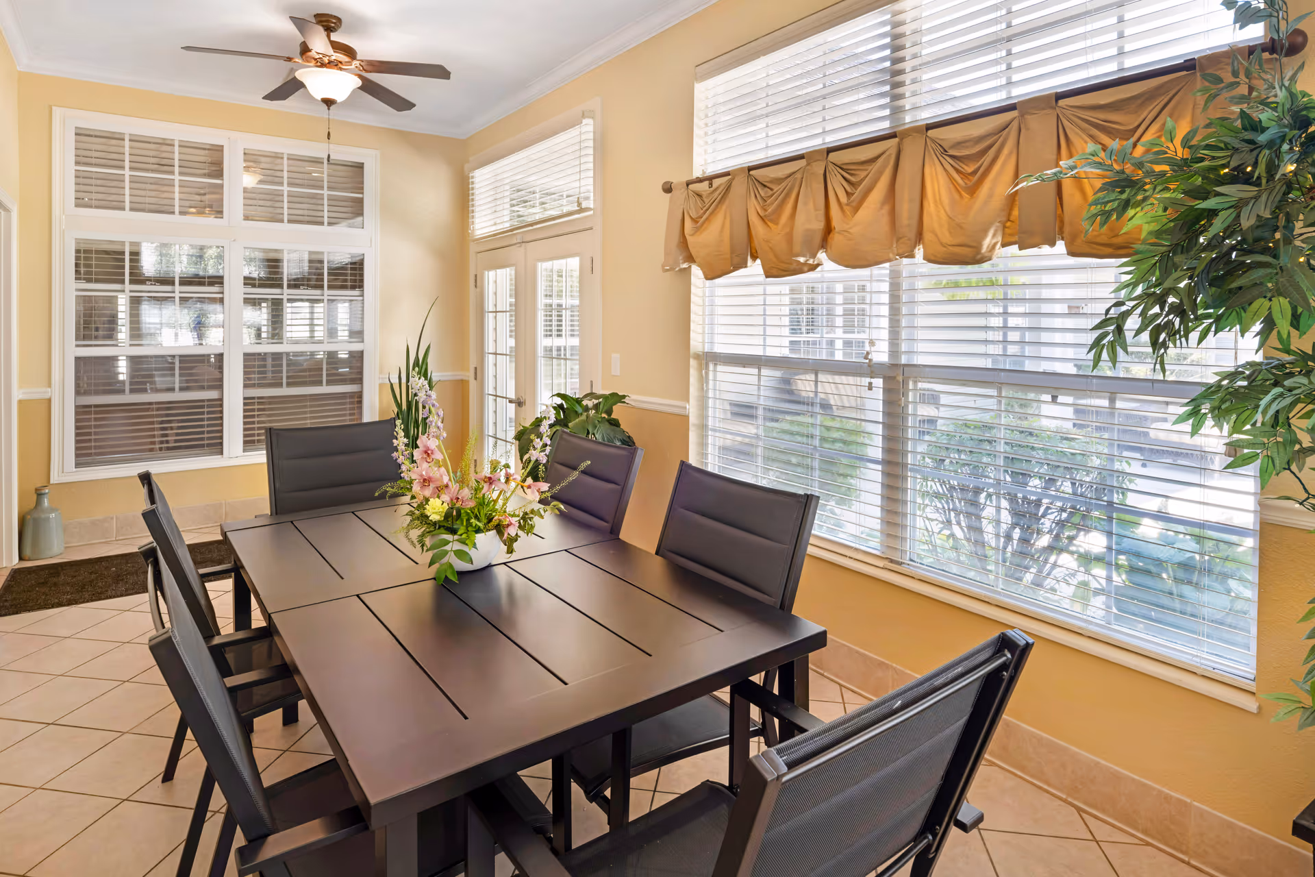 Sunlit dining room with a dark rectangular table and six chairs, a floral centerpiece, large windows with blinds and a valance, and a ceiling fan.