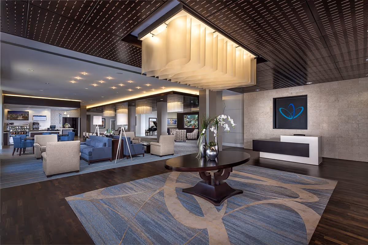 A spacious and modern senior living facility lobby with a large decorative ceiling light fixture, a round wooden table with vases and flowers in the center, comfortable seating areas with blue and beige chairs and sofas, a reception desk with a blue logo on the wall behind it, and a coffee station in the background.