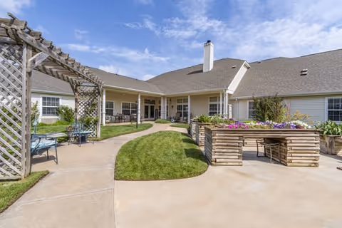 Outdoor patio area at Brookdale Enid featuring a paved walkway, green grass, wooden pergola with seating, raised garden beds with flowers, and a building with a covered porch in the background under a partly cloudy sky.