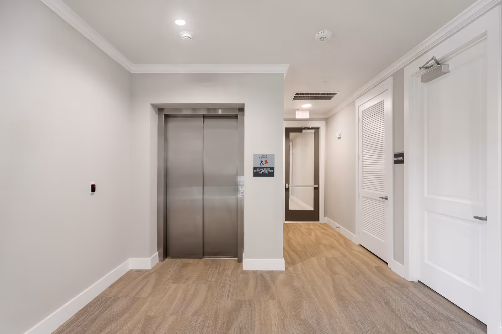 Interior hallway of a senior living facility with a closed stainless steel elevator door in the center, beige carpeted floor, light gray walls, and white doors on the right side. There is a sign next to the elevator indicating it is on level 2 and instructions for fire safety. A glass door with an exit sign above it is visible at the end of the hallway.