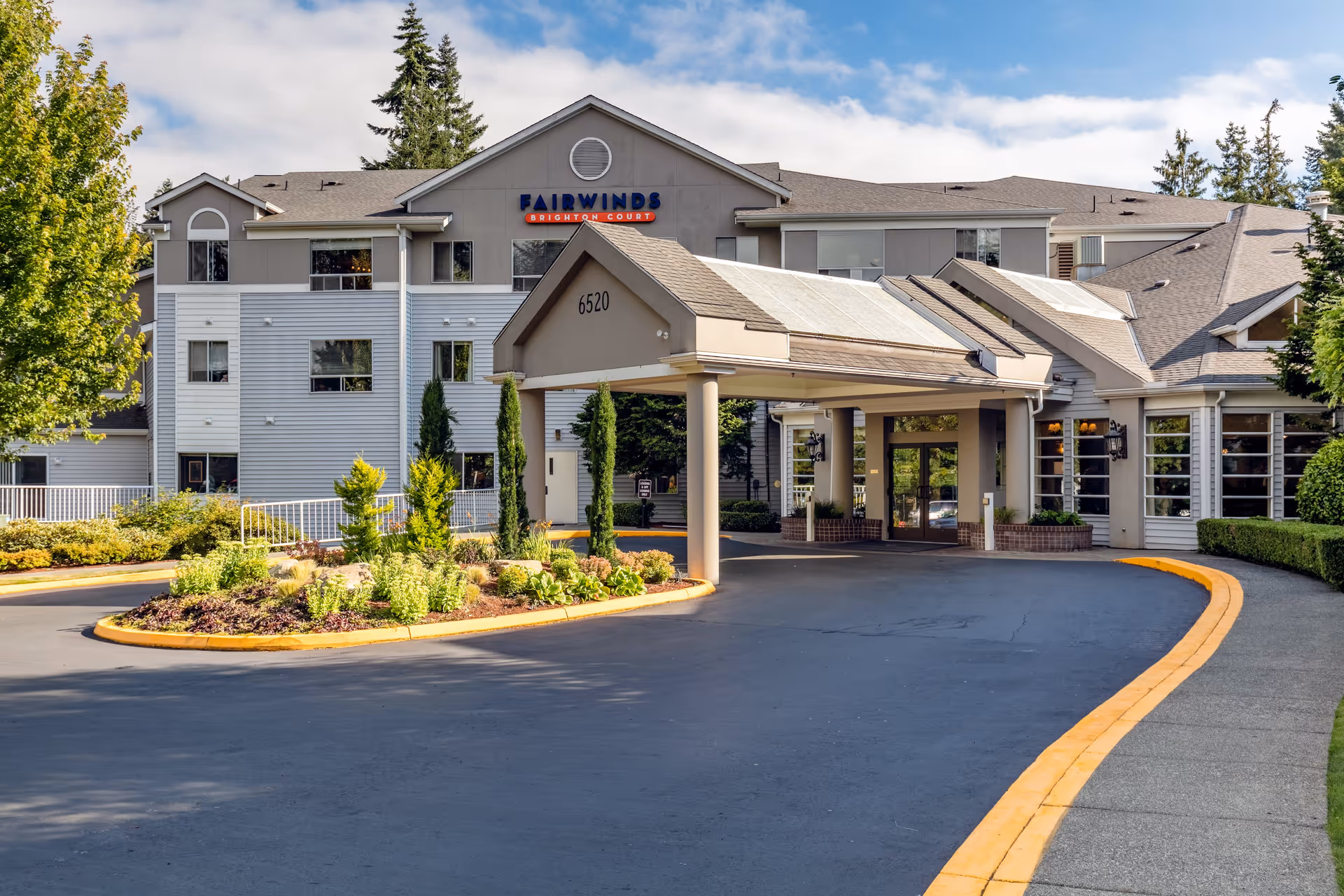 Exterior view of Fairwinds Brighton Court senior living facility showing the main entrance with a covered driveway, landscaped roundabout with shrubs and small trees, and a multi-story building with windows and a gray roof under a partly cloudy sky.