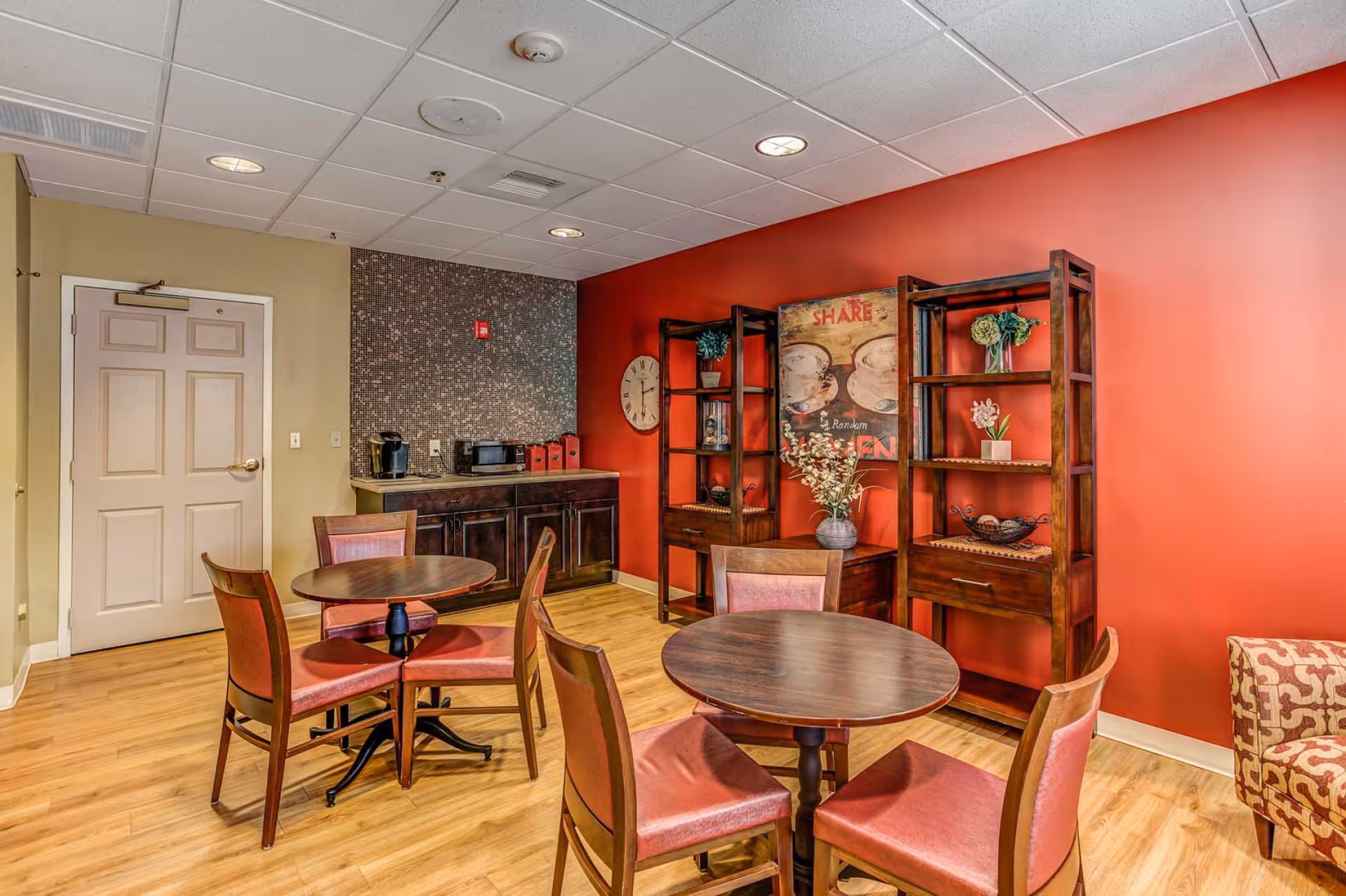 Small communal dining area with round tables and chairs, wooden shelving and a coffee station against a red accent wall.