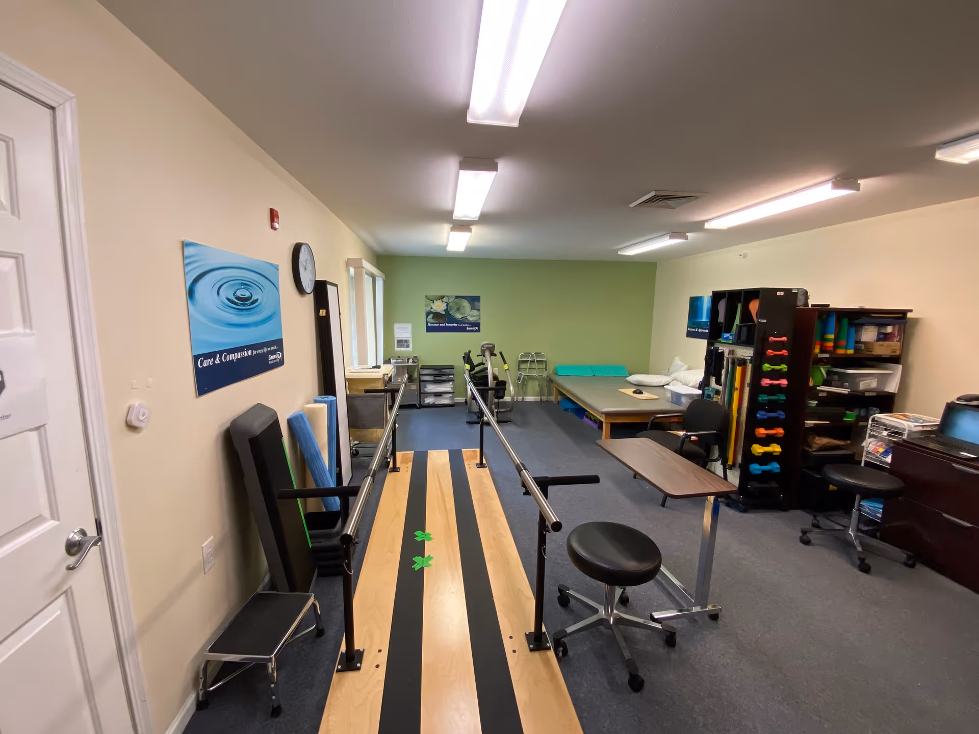 Interior view of a therapy or rehabilitation room with parallel bars for walking exercises, a padded therapy table, exercise equipment including small dumbbells, and chairs. The walls have motivational posters and the room is well-lit with overhead fluorescent lights.
