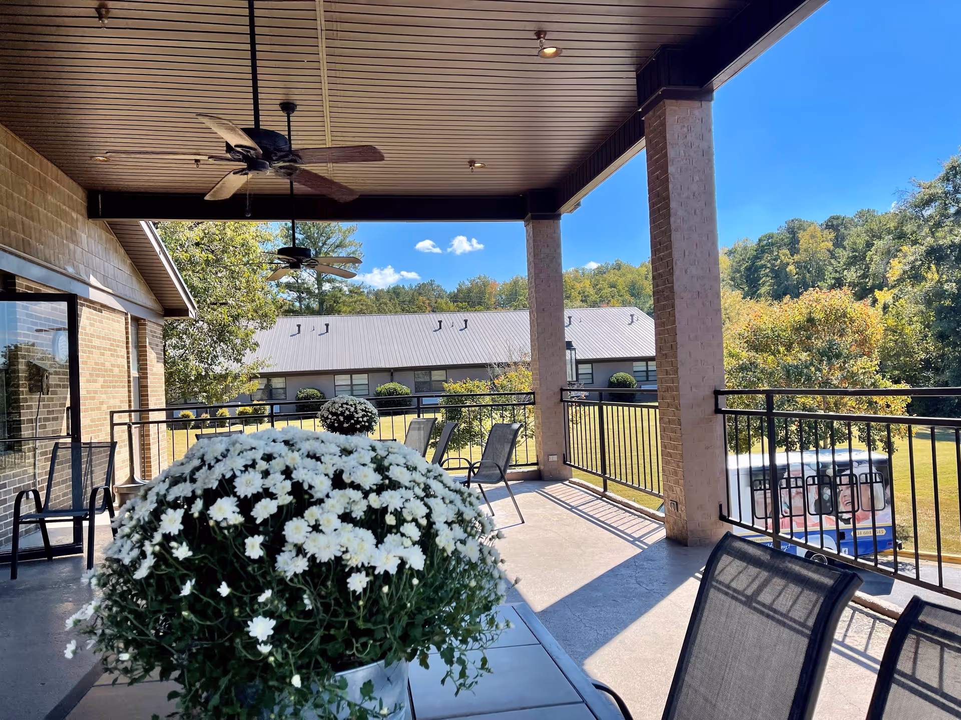 Covered outdoor patio area with ceiling fans, metal railing, and patio furniture including chairs and tables with potted white flowers. In the background, there is a grassy area with trees and a building with a metal roof under a clear blue sky.