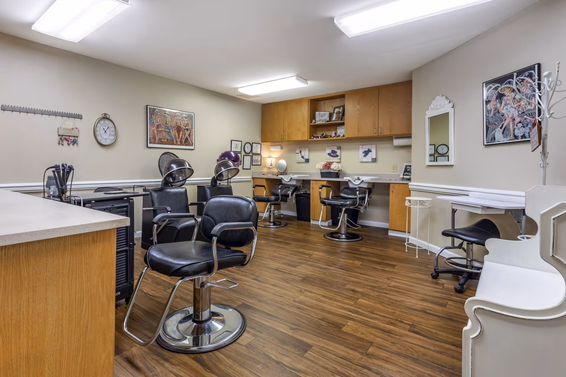 Interior of a hair salon room with wooden flooring, black salon chairs, hair dryers, sinks for washing hair, wooden cabinets, wall art, a clock, and a white coat rack.